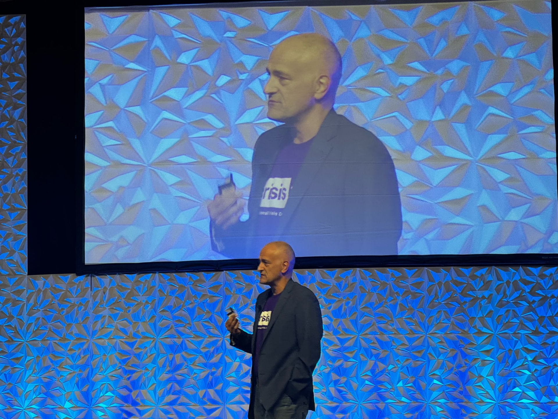 Eric Arzubi, MD, delivering a keynote on stage at CrisisCon26, presentation remote raised, with a live camera feed of him on the screen behind.